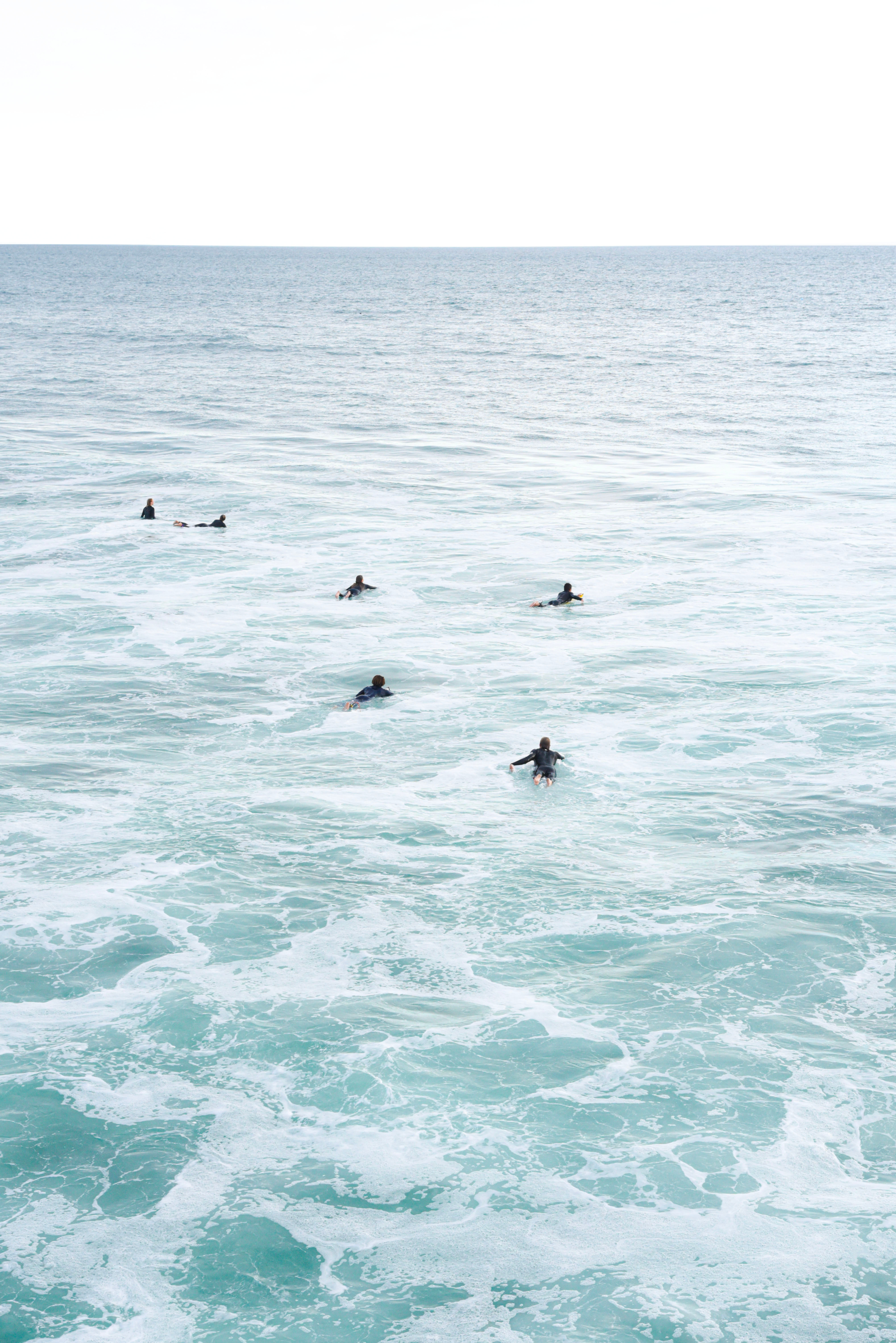 A color photograph of surfers paddling out in the blue-green waters speckled with the residual froth of a wave in Oceanside.