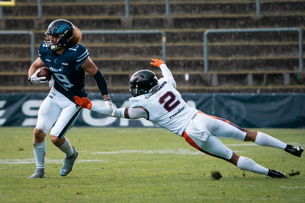American football player in blue runs with the ball as a white-clad defender dives to tackle him on a rainy field.