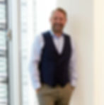 Smiling man in blue vest and shirt leans against a wall in a bright office with large windows, exuding a relaxed and confident demeanor.