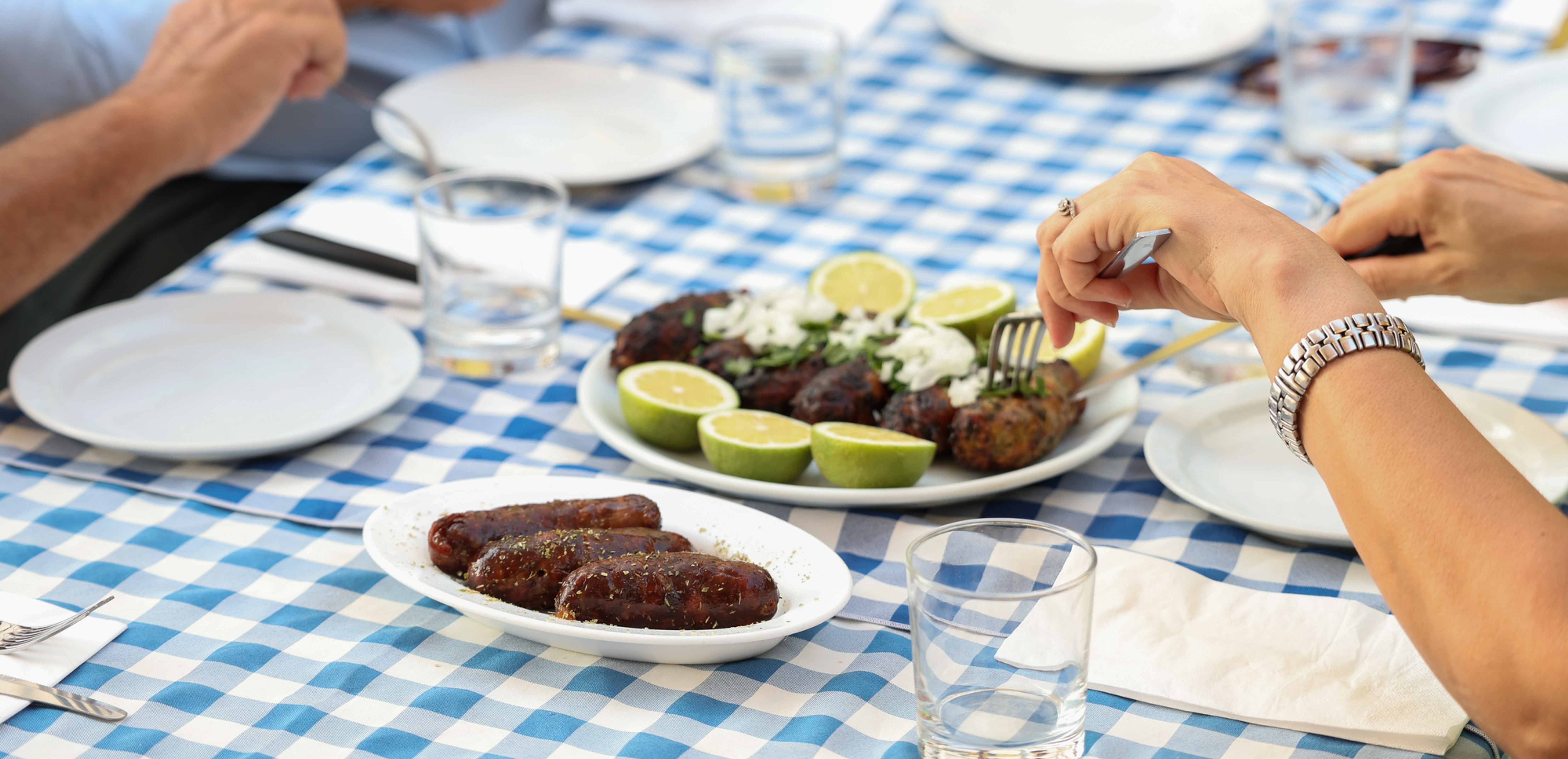 Hands of several people share grilled food and limes at a blue-checkered outdoor table with plates and glasses.