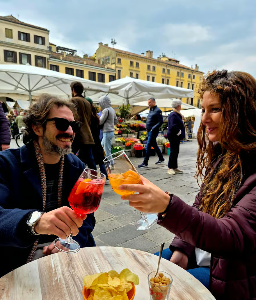 Man and woman smile and toast with red and orange aperitifs at a piazza cafรฉ, with market umbrellas, flowers, and historic buildings behind them. FOODAQUEST Padova food tour