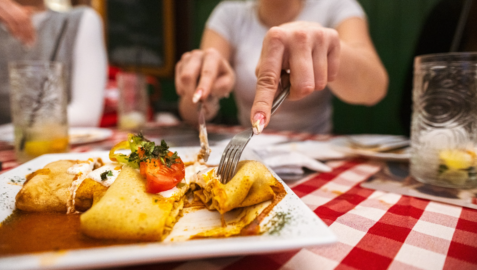 Two women at a restaurant cut into stuffed crepes on a plate atop a red checkered tablecloth.