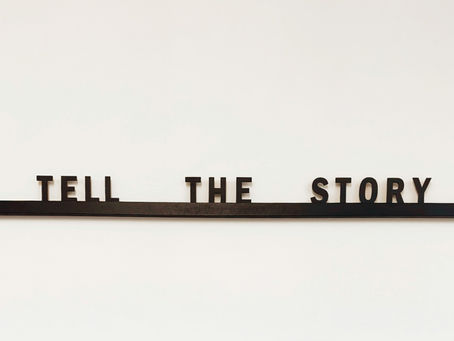 Black letters spelling "TELL THE STORY" on a white wall. The letters rest on a narrow horizontal black shelf, creating a minimalist look.