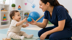 Cuidadora de azul brinca com bebê sentado em sala terapêutica, trocando bola azul; brinquedos coloridos e clima alegre.