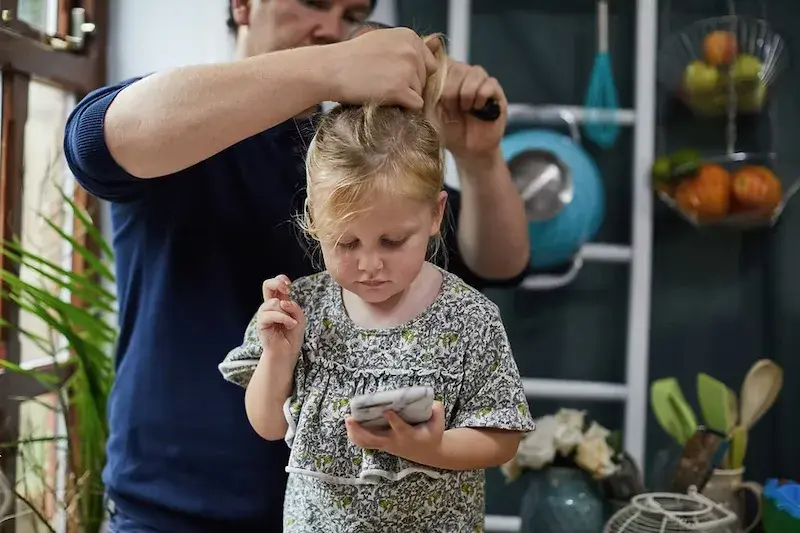 father tying up daughters hair while she looks at phone