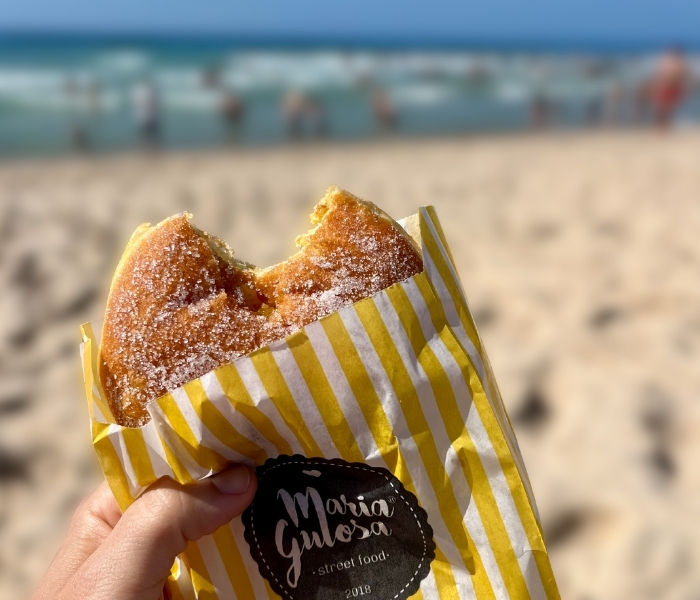 A bola de berlin with a bite taken out of it sticks out from a white and yellow striped bag, with a blurred beach in the background