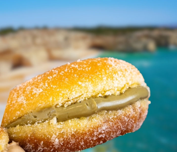 A bola de berlim filled with green pistachio filling against a blurred background of a cliffside beach in Portugal