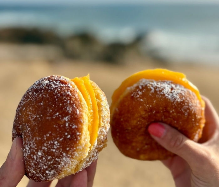 Two hands hold up traditional custard bola be berlims with a Portuguese beach blurred in the background