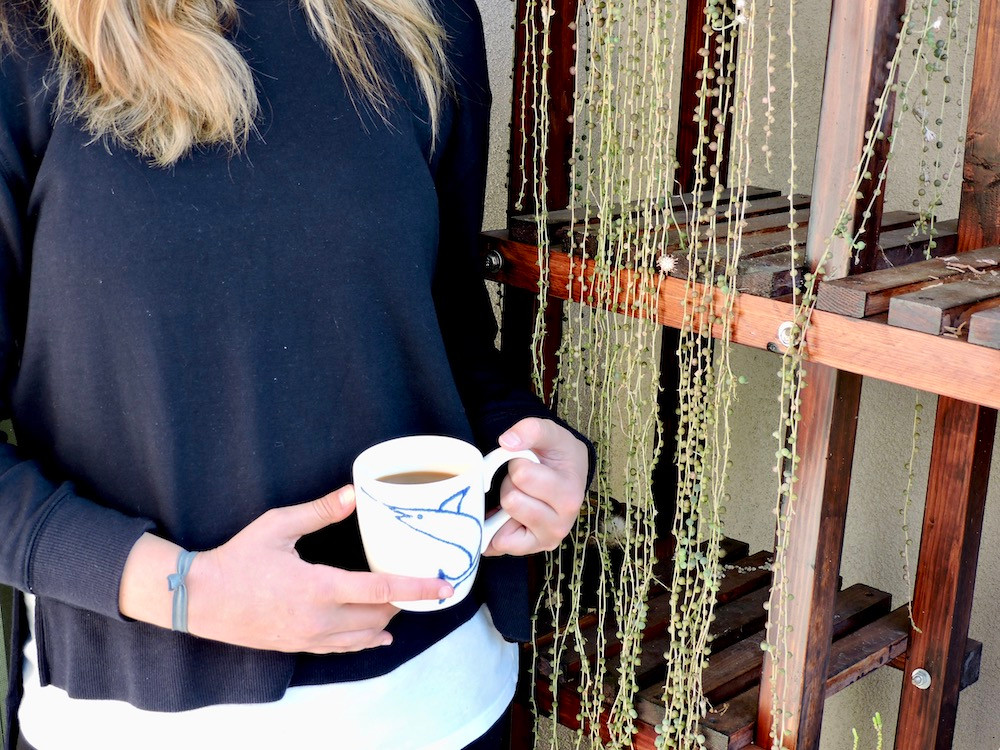 Girl stands near plant stand holding cup of Saudade Coffee