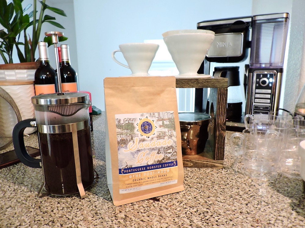 A bag of Saudade Coffee sits near a french press, pour over coffee maker, and a coffee pot