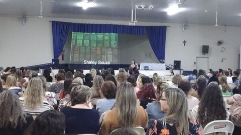 Foto da autora Shirley Souza palestrando para professores em um auditório.