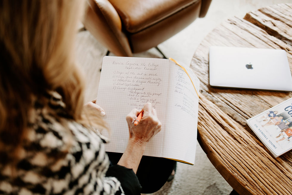Woman writing in a notebook at a wooden table with a laptop and book. Setting appears cozy and focused, with neutral tones.