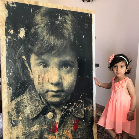 Mina at home with her wooden portrait. She had just added her tiny hand and foot prints to the bottom of the image in red and gold paint.
Additional story from the family: "My name is Mina I am 2 years and 2 months old. I am the younger sister of two brothers. My parents and grandparents are all from Kurdistan but I was born in Ipswich, UK. I am a 'Daddy's Girl'."