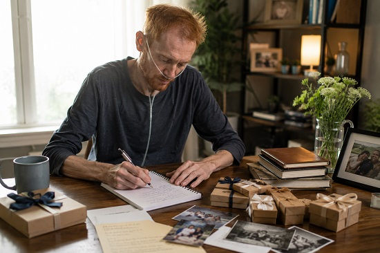 A very thin red-haired man sits at a wooden table near a bright window, writing carefully in a notebook as he documents the story of his life. An oxygen cannula rests beneath his nose, and his frail appearance suggests serious illness. Spread across the table are family photographs, keepsake boxes tied with ribbons, journals, and personal mementos that appear to help him remember different chapters of his past. Several completed gift boxes sit beside others still waiting to be finished. Soft natural daylight fills the room, creating a reflective and intimate atmosphere as he records memories, experiences, and family stories for the people he loves.