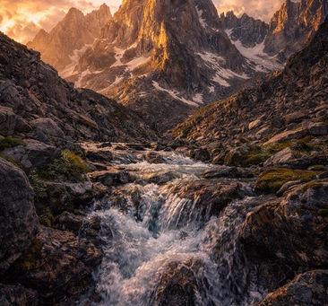 Towering jagged mountain peak rising above a rocky valley with a rushing stream in the foreground, illuminated by warm natural daylight beneath dramatic clouds, symbolizing divine majesty.