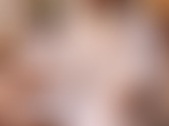 An elderly Sikh man wearing a white turban and traditional white garments sits cross-legged on a patterned carpet inside a gurdwara, his hands resting open on his knees in a posture of meditation. His eyes are gently lowered, and his long white beard frames a calm, inward-focused expression. In the softly blurred background, another Sikh man sits in prayer beneath stone arches, as warm natural daylight fills the sacred space with stillness and reverence.