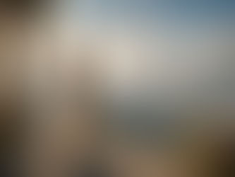 A woman in flowing cream robes stands on an ancient stone terrace overlooking a vast mountain landscape beneath a bright daytime sky. Around her, crystal-clear floating spheres reflect galaxies, cities, and distant worlds, suggesting that awareness may extend beyond any single place. Warm natural sunlight, timeless architecture, and expansive scenery create a contemplative image exploring the question of whether consciousness is local or infinite.