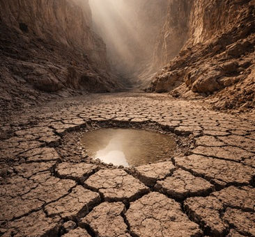 Sunlit desert canyon with cracked earth in the foreground and a small reflective pool at the center, warm golden daylight illuminating textured rock walls, symbolizing contraction and containment in nature.