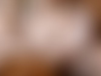 An elderly Sikh man wearing a white turban and traditional white garments sits cross-legged inside a gurdwara, playing a wooden harmonium as he sings in devotion. His hands rest gently on the keys, his long white beard framing a focused, prayerful expression. The instrument sits on a patterned rug beside a small bowl of flowers and prayer beads, while stone arches and soft daylight create a calm, sacred atmosphere of kirtan and reverent sound.