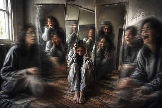 A distressed person sits alone on the floor with their hands pressed tightly against their head while multiple distorted reflections and blurred figures appear around them in tall mirrors. The ordinary room contrasts with the unsettling visual distortions, symbolizing the confusion, fear, and altered perception that many people experiencing psychosis describe, while conveying the profound isolation and emotional intensity of the experience.