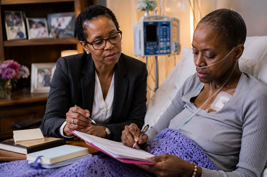 A seriously ill Black woman sits upright in a hospital bed, wrapped in a blanket and wearing a nasal oxygen cannula, as she carefully writes a personal letter. Beside her, a professionally dressed woman reviews documents and offers guidance, creating the impression of a thoughtful discussion about values, wisdom, and the legacy she wishes to leave behind. Family photographs and notebooks sit nearby, while medical equipment in the background underscores the reality of her illness. The scene captures a deeply meaningful moment of reflection as she creates an ethical will and legacy letter, passing on life lessons, memories, and personal beliefs to future generations