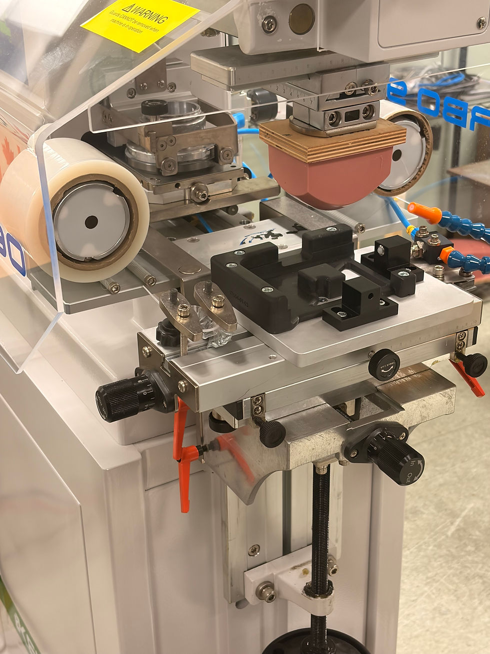 Close-up of an industrial machine with rollers, clamps, and gauges under a clear shield, plus a yellow WARNING label.