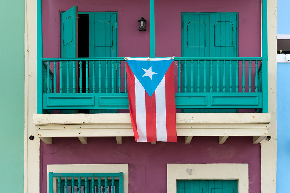 A Puerto Rican flag hangs from a teal balcony on a vibrant pink wall. Open shutters and closed doors add charm to the colorful building.