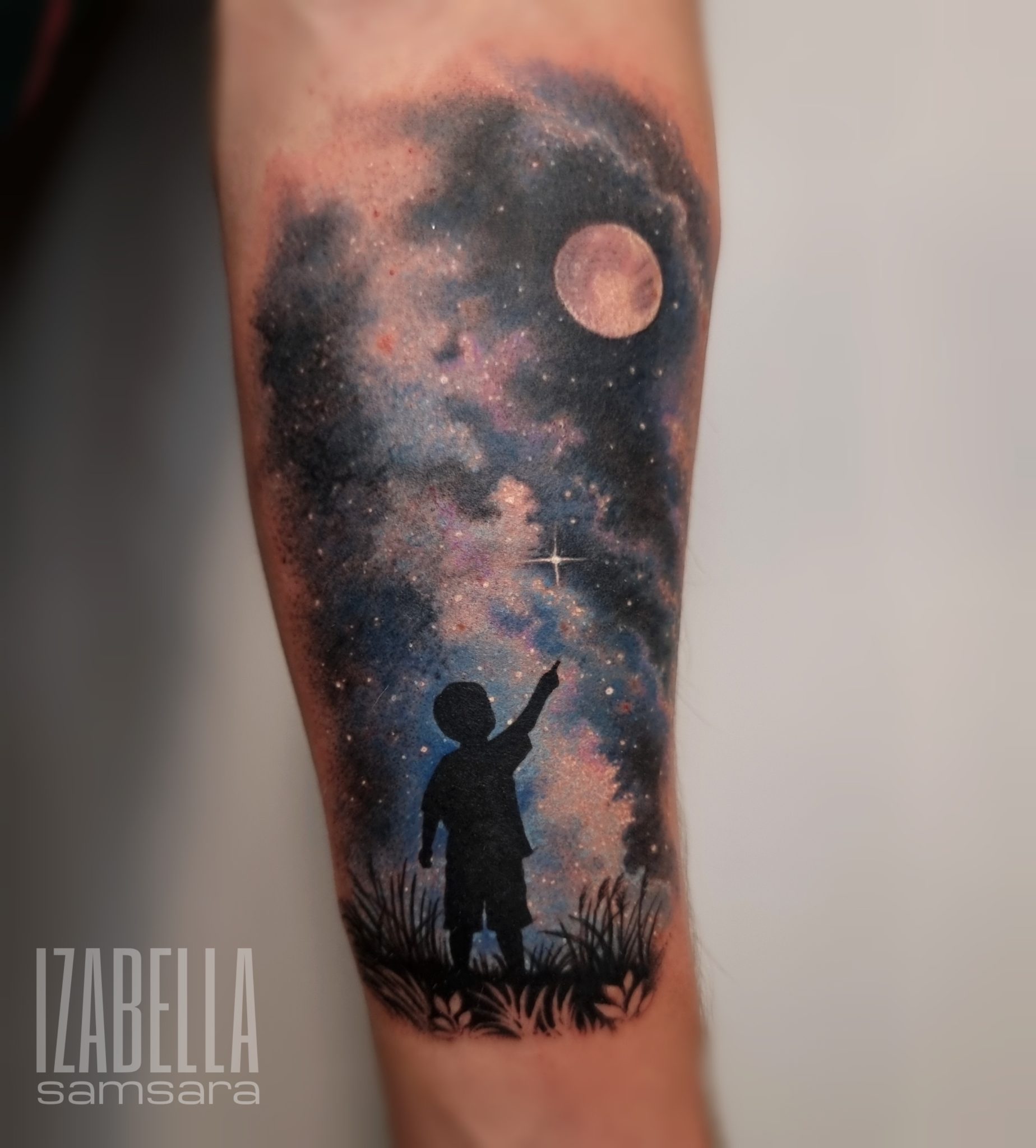 A vibrant tattoo depicting a child silhouette standing on grass and pointing at a starry night sky, with a large moon and colorful cosmic clouds in the background.