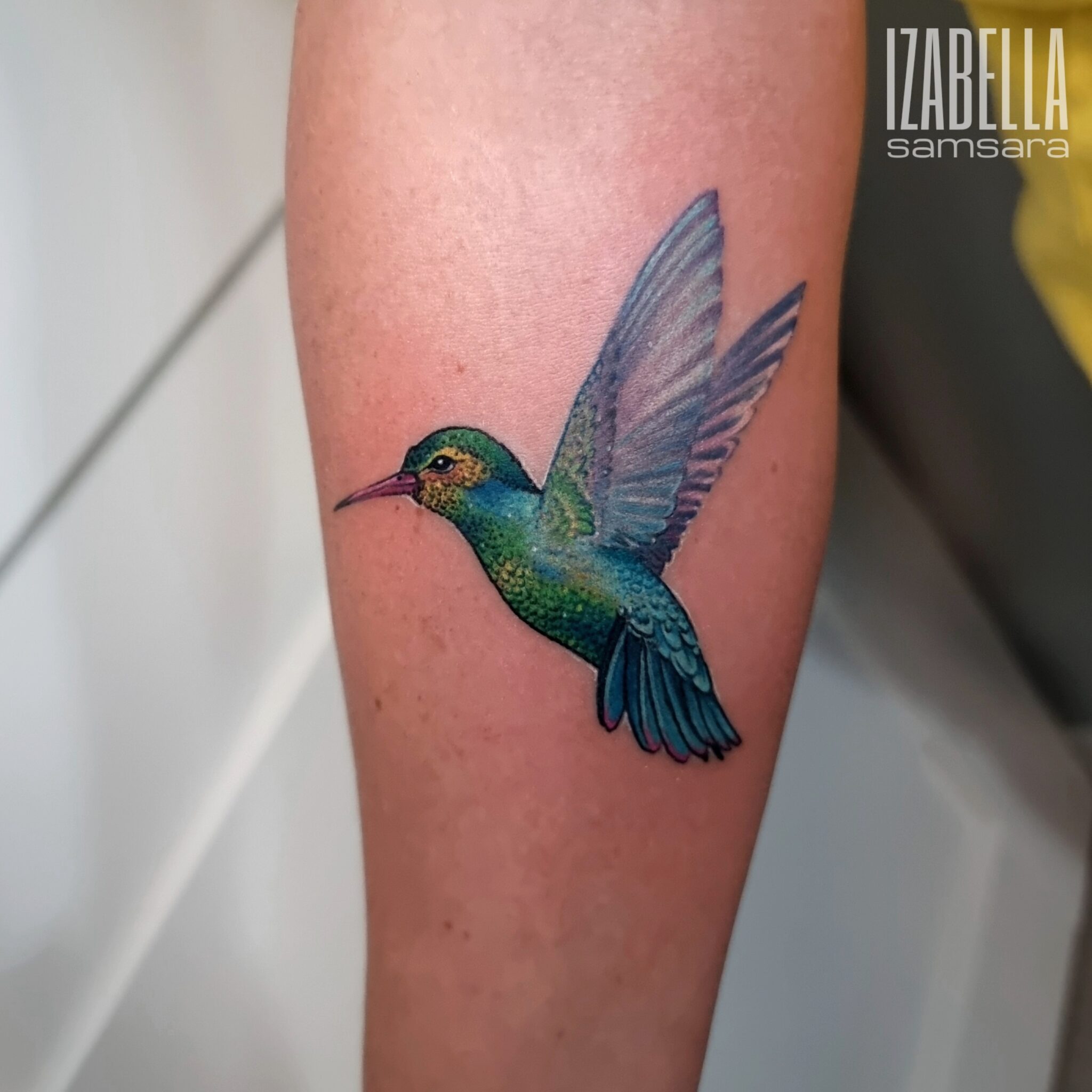A vibrant tattoo of a hummingbird in flight, showcasing intricate detailing and bright colors, including shades of green and blue. The hummingbird's wings are spread wide and appear graceful, set against a subtle background.