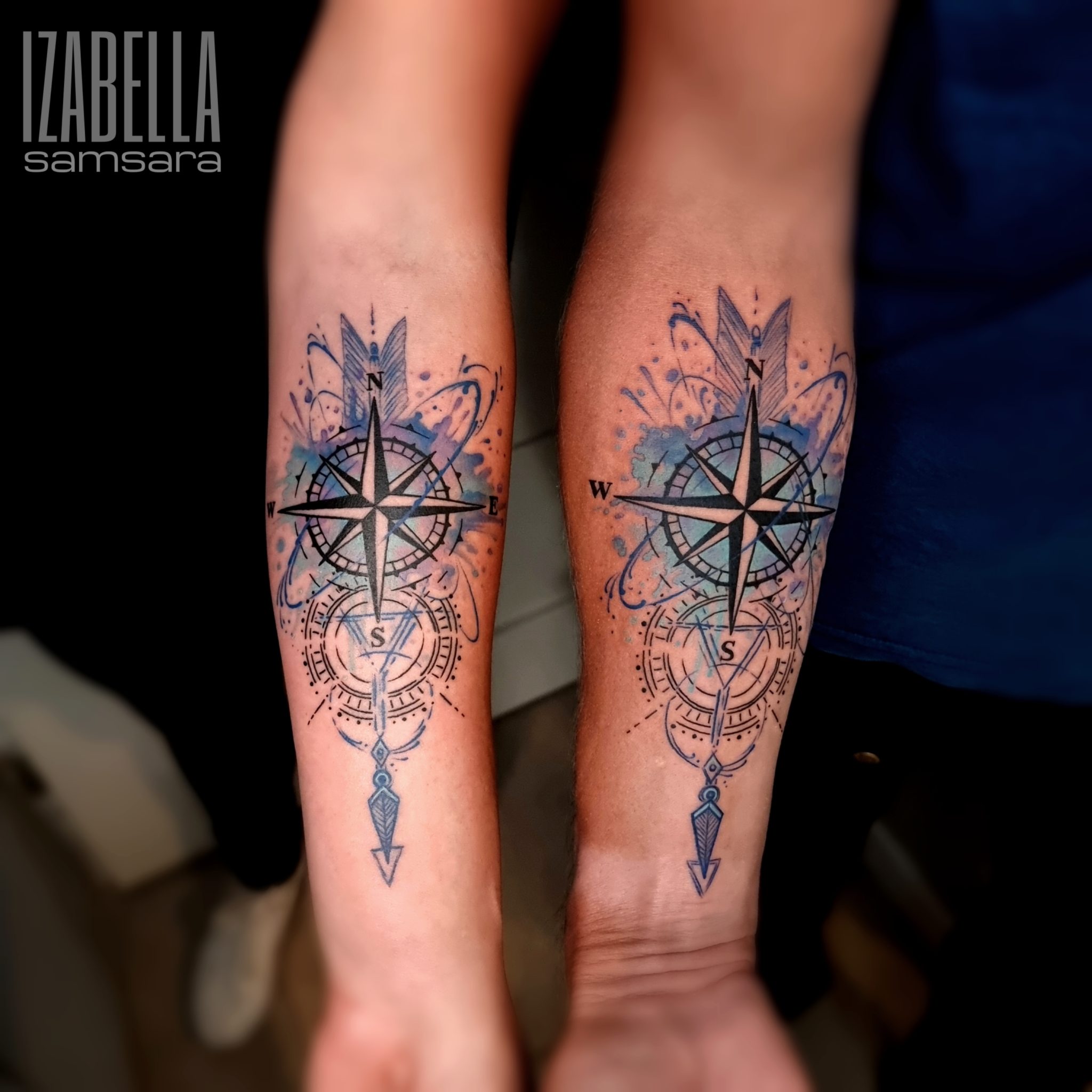 A vibrant tattoo of a compass, featuring intricate details and a watercolor background in shades of blue. The compass has directional markers (N, E, S, W) clearly visible, surrounded by decorative swirls and patterns.