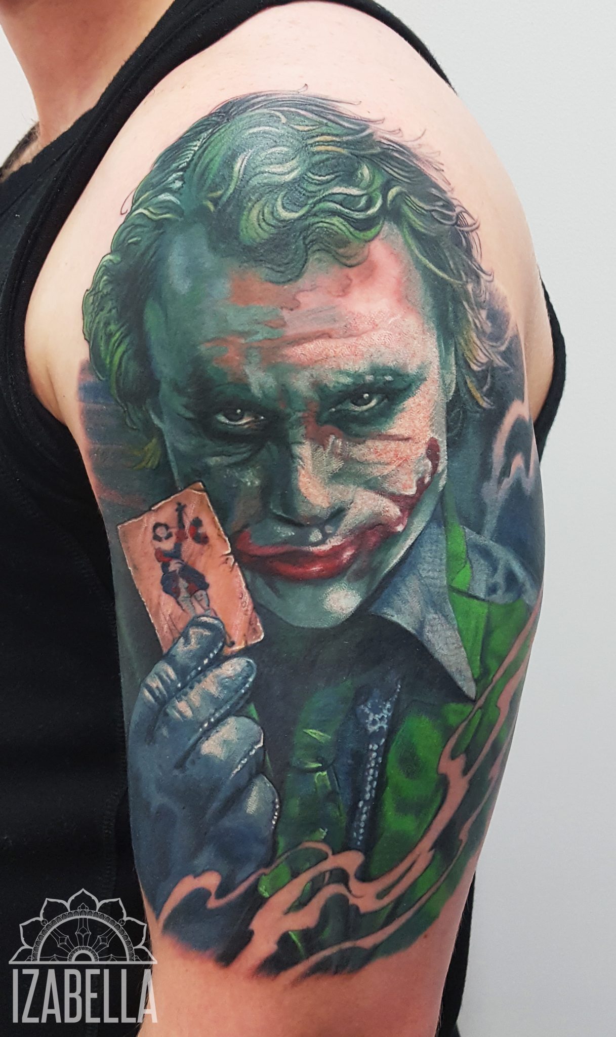 A vivid tattoo of the Joker, featuring detailed facial features with green skin, red lipstick, and a sinister grin. The character holds a playing card with a painted image of a woman and is dressed in a green suit with a white collar. The background includes swirling colors, enhancing the dynamic and dramatic effect of the tattoo.
