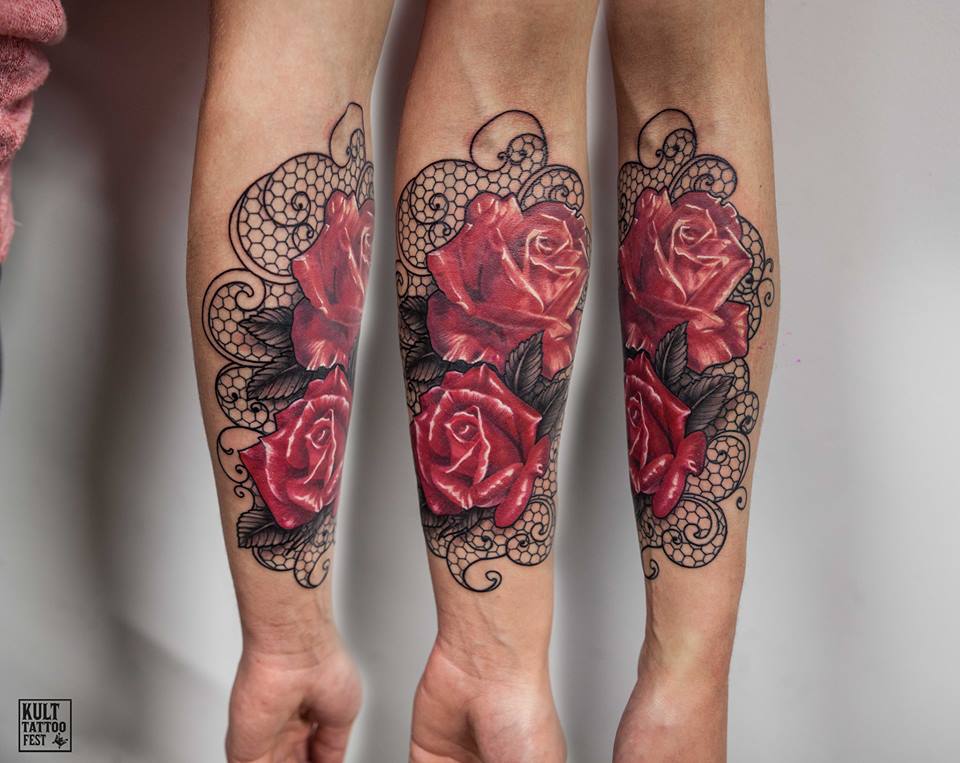 A vibrant tattoo featuring detailed red roses with dark green leaves, surrounded by intricate black lace designs. The tattoo covers both forearms, creating a symmetrical look, showcasing the beauty and elegance of floral artistry.