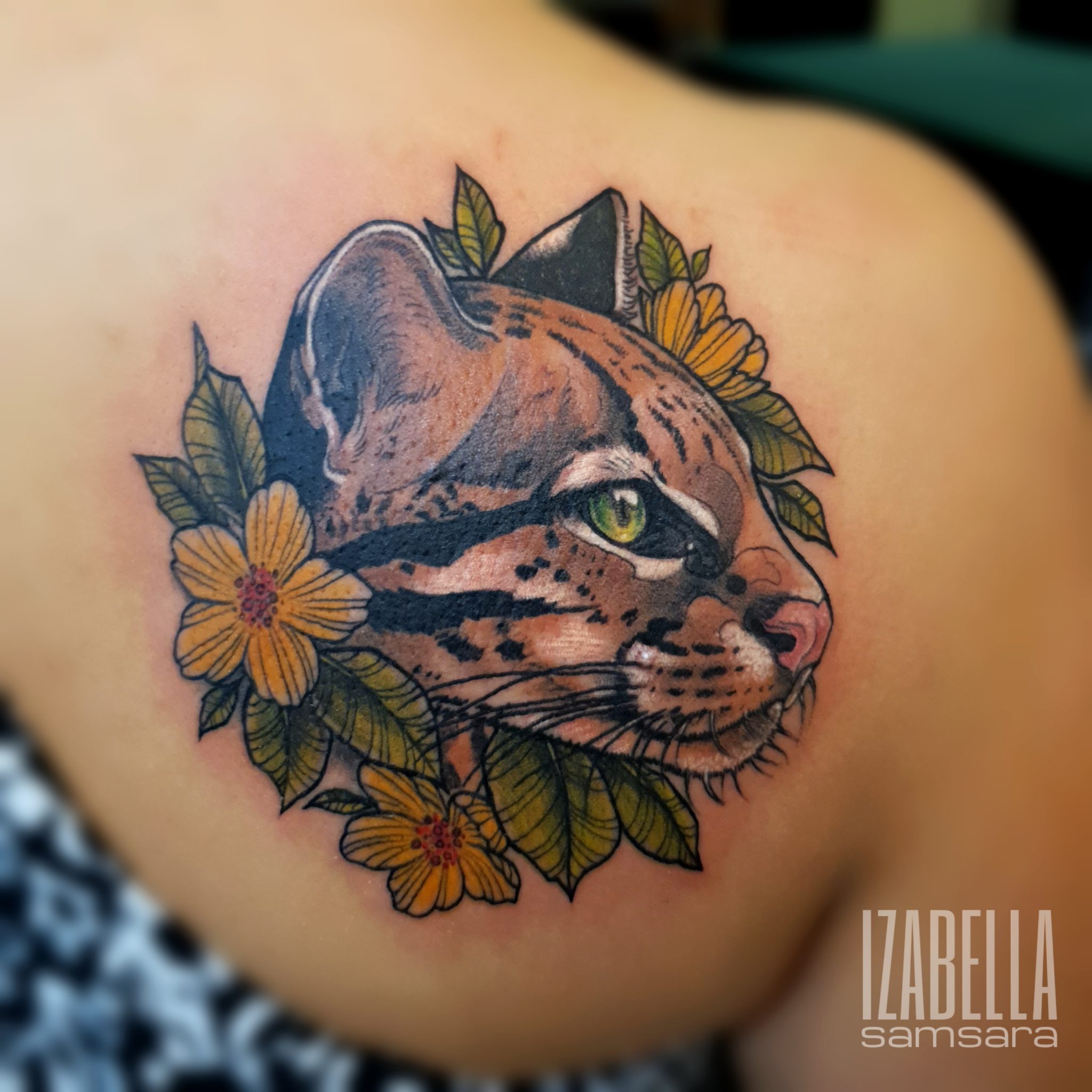 A beautifully detailed tattoo of a wild cat's head surrounded by vibrant yellow flowers and green leaves. The cat features striking green eyes and realistic fur patterns, showcasing a blend of brown, black, and hints of gray. The floral elements add a touch of nature, enhancing the artwork's appeal.