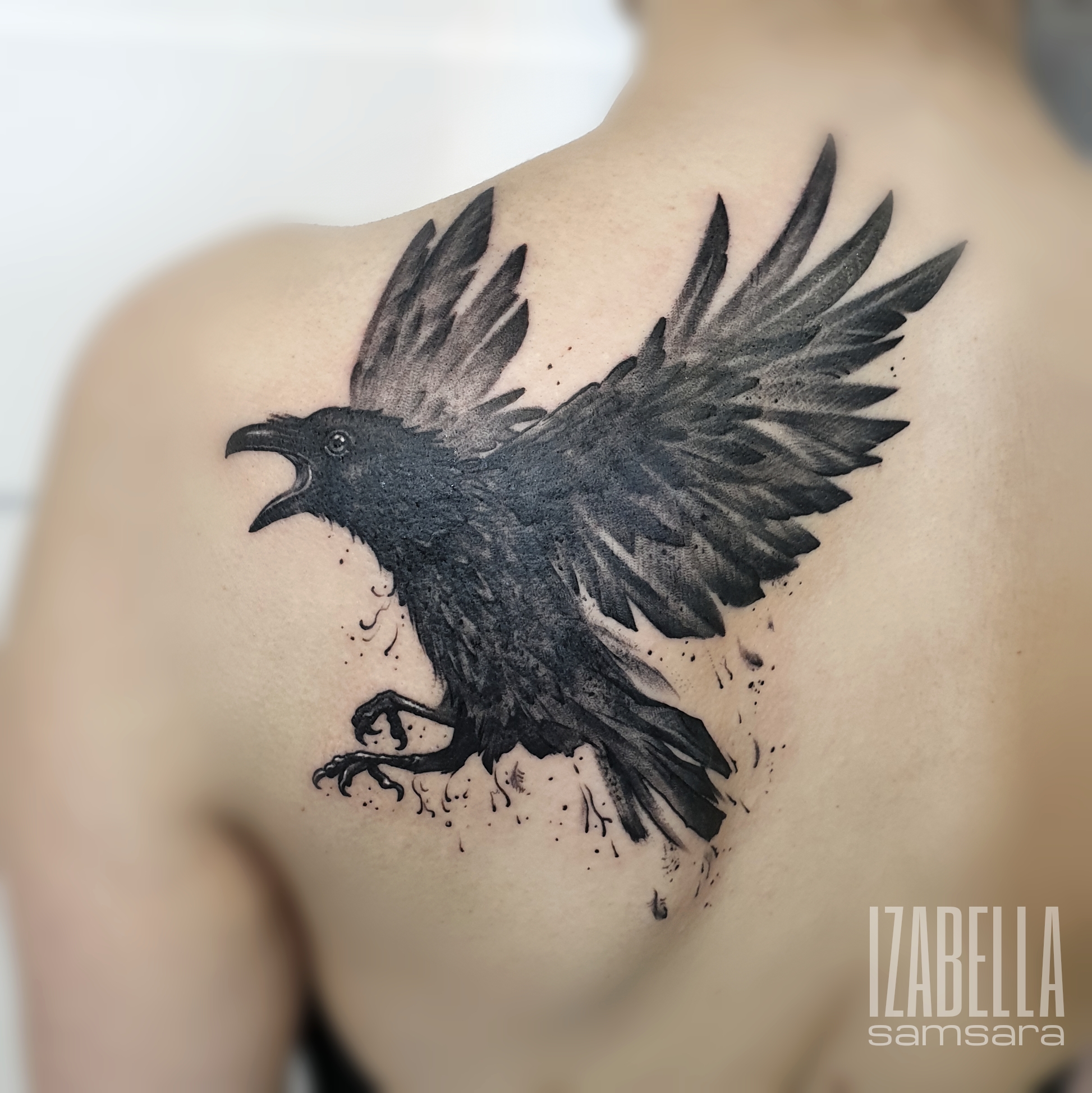 A striking black and grey tattoo of a raven in mid-flight, located on the upper back. The raven is depicted with its wings outstretched and mouth open, creating an effect of motion. The design includes intricate feather details, along with splashes of ink that enhance its dynamic appearance.