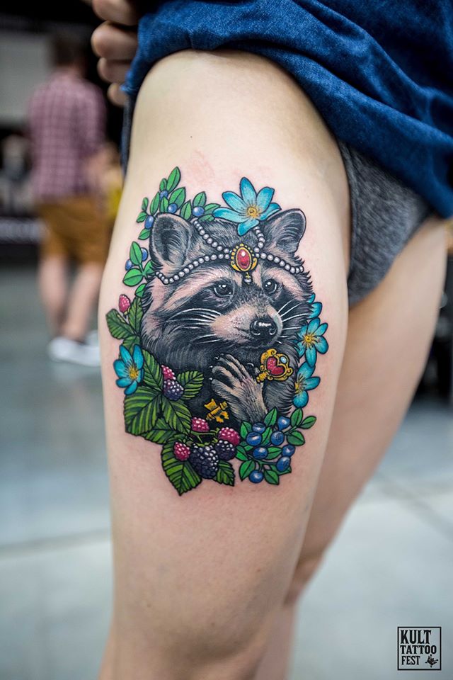A colorful tattoo featuring a raccoon adorned with a jeweled headpiece and surrounded by vibrant flowers and greenery. The raccoon has intricate details, showcasing its playful expression and whiskers, while the floral elements include blue flowers and lush green leaves, creating a lively and whimsical design that highlights the beauty of nature.