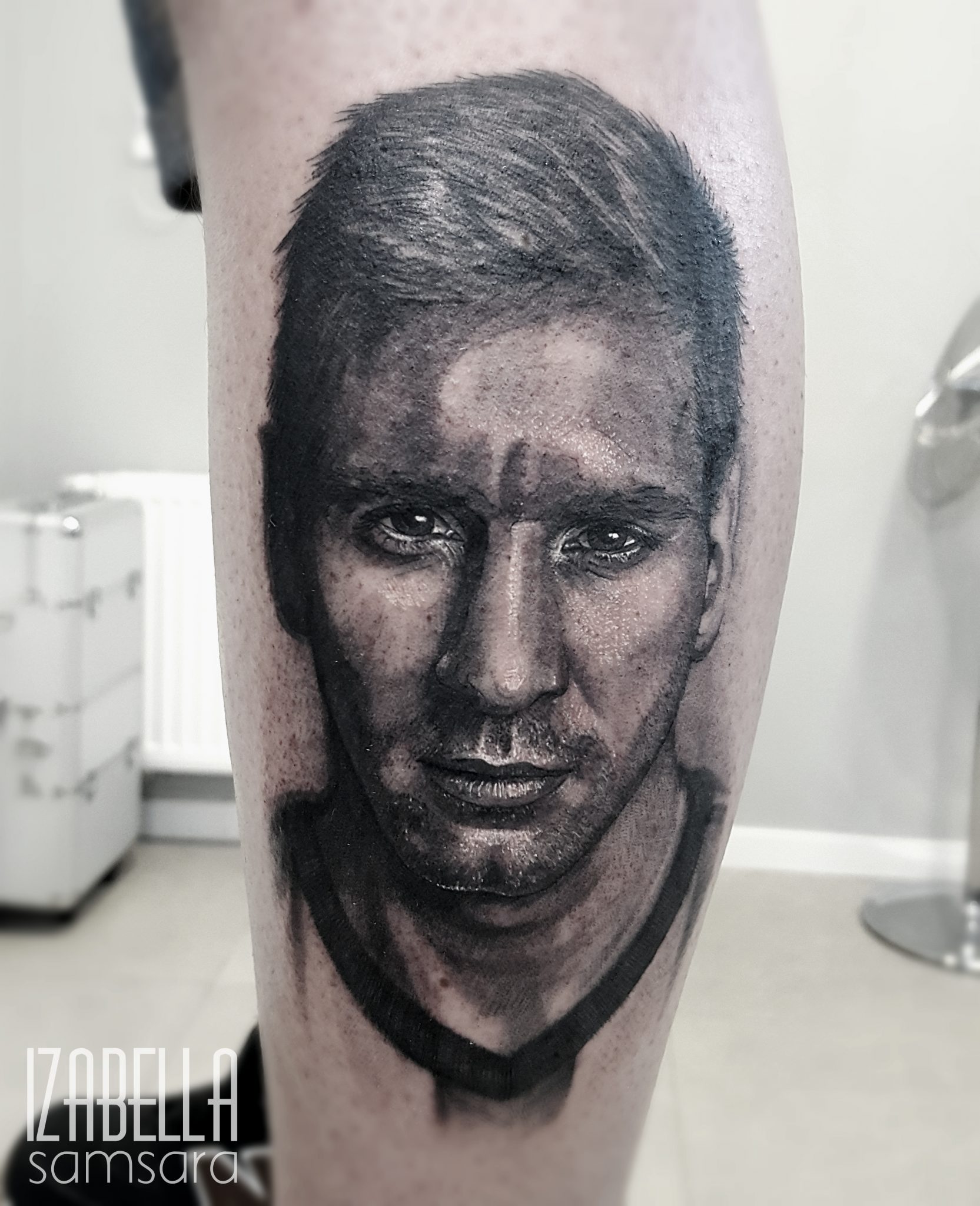 A highly detailed black and white tattoo of a male face, showcasing realistic features, intense expression, and intricately shaded hair. The image captures fine details in the eyes and skin texture, providing a lifelike representation.