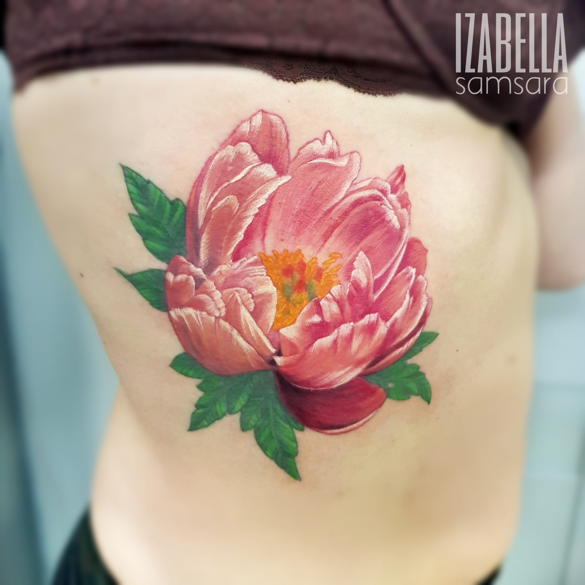 A vibrant tattoo of a large pink peony flower with green leaves, located on the side of the torso. The peony features detailed petals with shades of pink, highlighted with touches of yellow and orange at the center. This tattoo combines realism and color, showcasing the beauty of floral art.