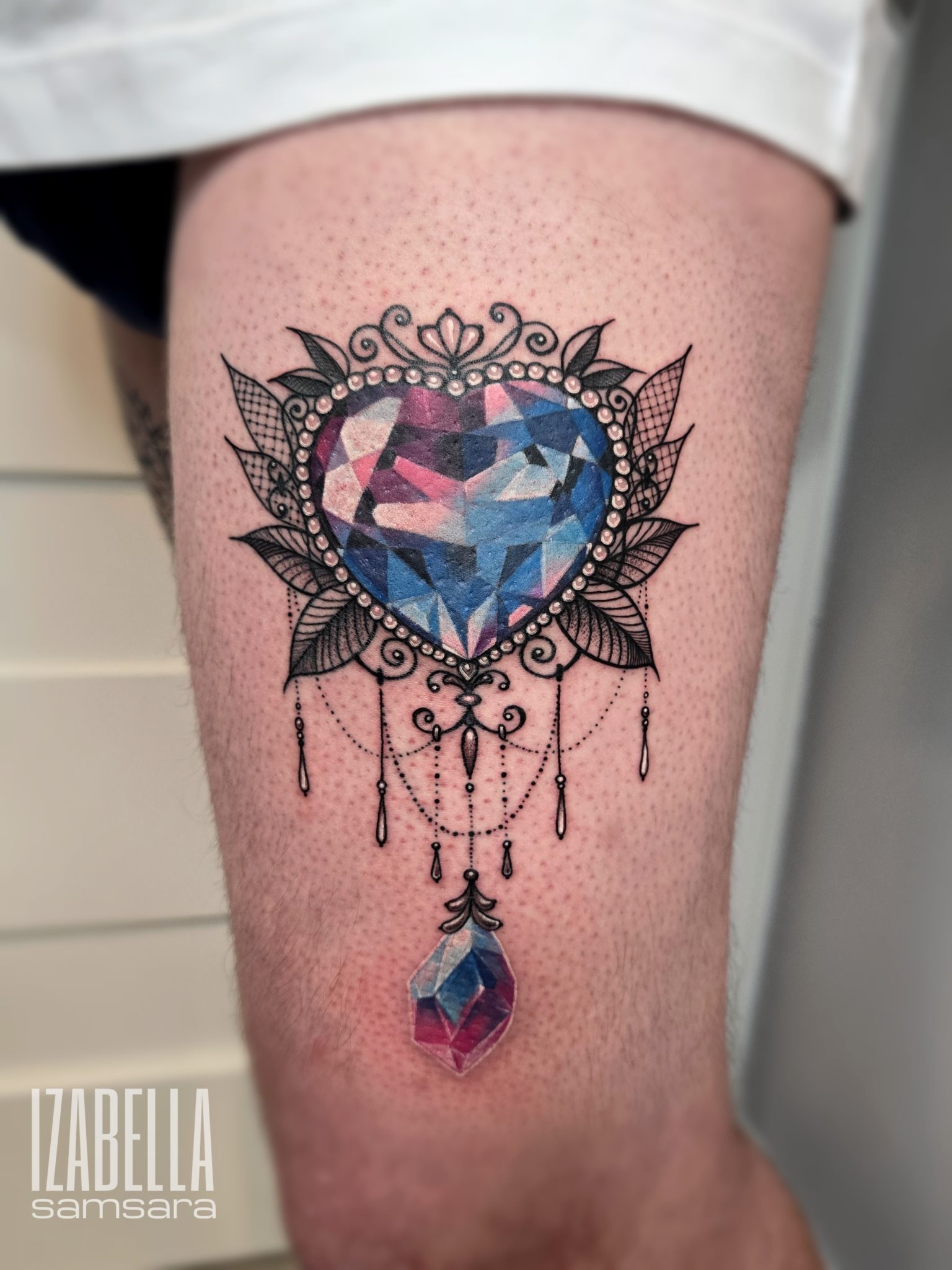 A vibrant tattoo featuring a large heart-shaped gemstone in shades of blue and pink, surrounded by intricate black lacework and decorative elements including pearls and leaves. Below the heart, there are elegant chains adorned with small droplets, leading down to a smaller crystal hanging at the bottom, all set on a light skin tone background.