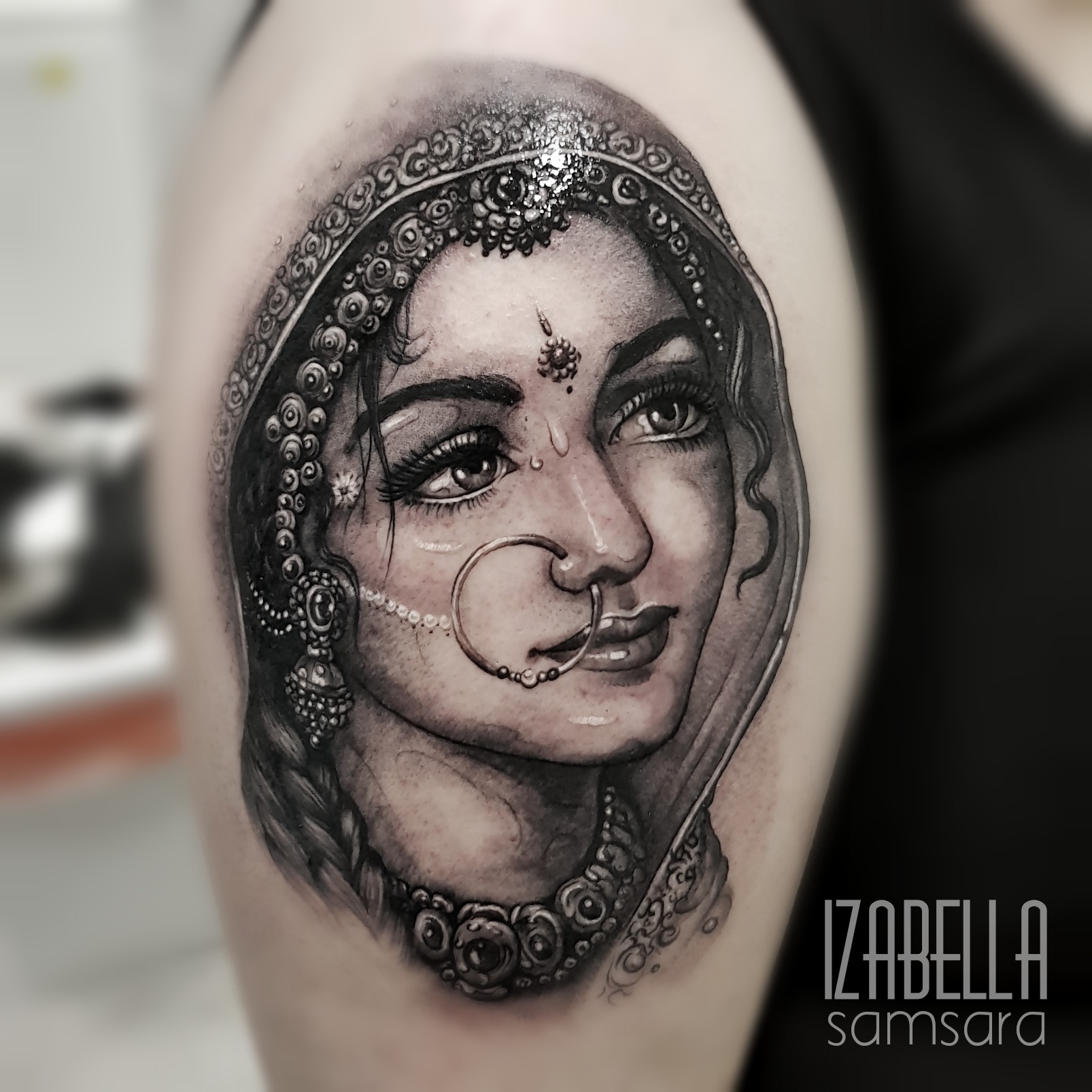 A realistic black and gray tattoo depicting a young woman wearing traditional jewelry, with intricate detailing on her face and a large nose ring. She has long, braided hair and is adorned with decorative forehead jewelry. The tattoo features soft shading and fine lines, showcasing the beauty of cultural adornments.