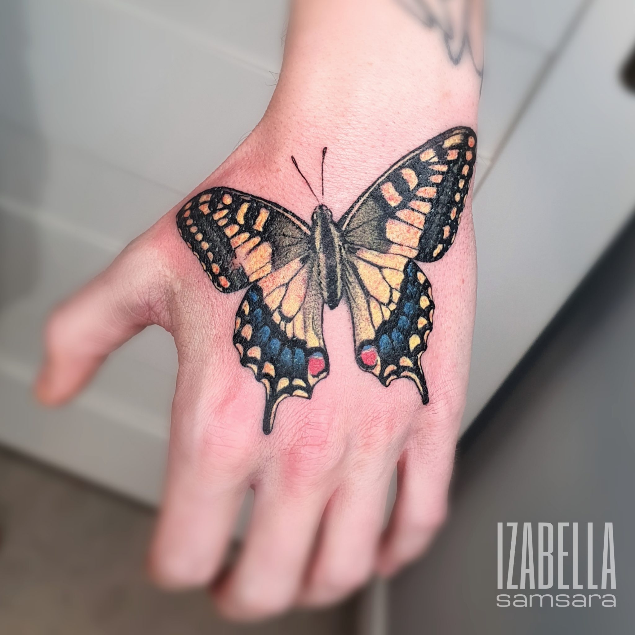 A vibrant hand tattoo featuring a detailed butterfly with intricate patterns. The butterfly is colored in shades of orange, blue, and black, showcasing its wings open wide. This tattoo is positioned on the back of the hand, highlighting the design against the skin.