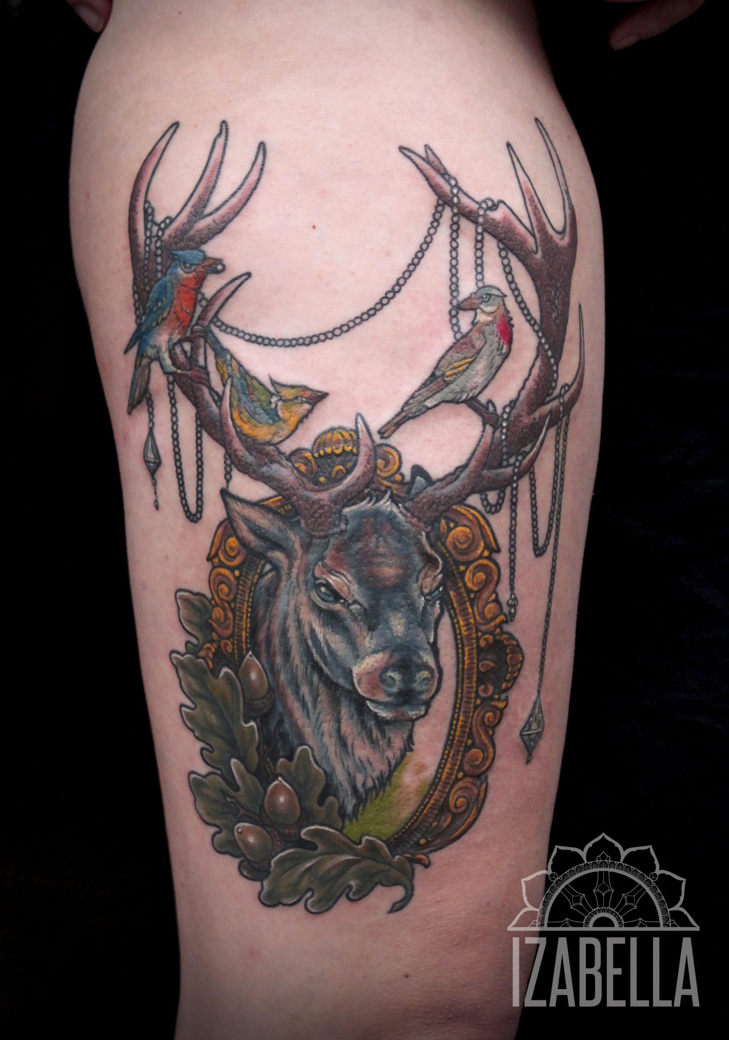 A vibrant tattoo featuring a detailed stag's head surrounded by ornate golden frames, adorned with hanging chains and colorful birds perched on its antlers. The design includes elements of nature like oak leaves and acorns, showcasing an artistic mix of wildlife and decorative motifs.
