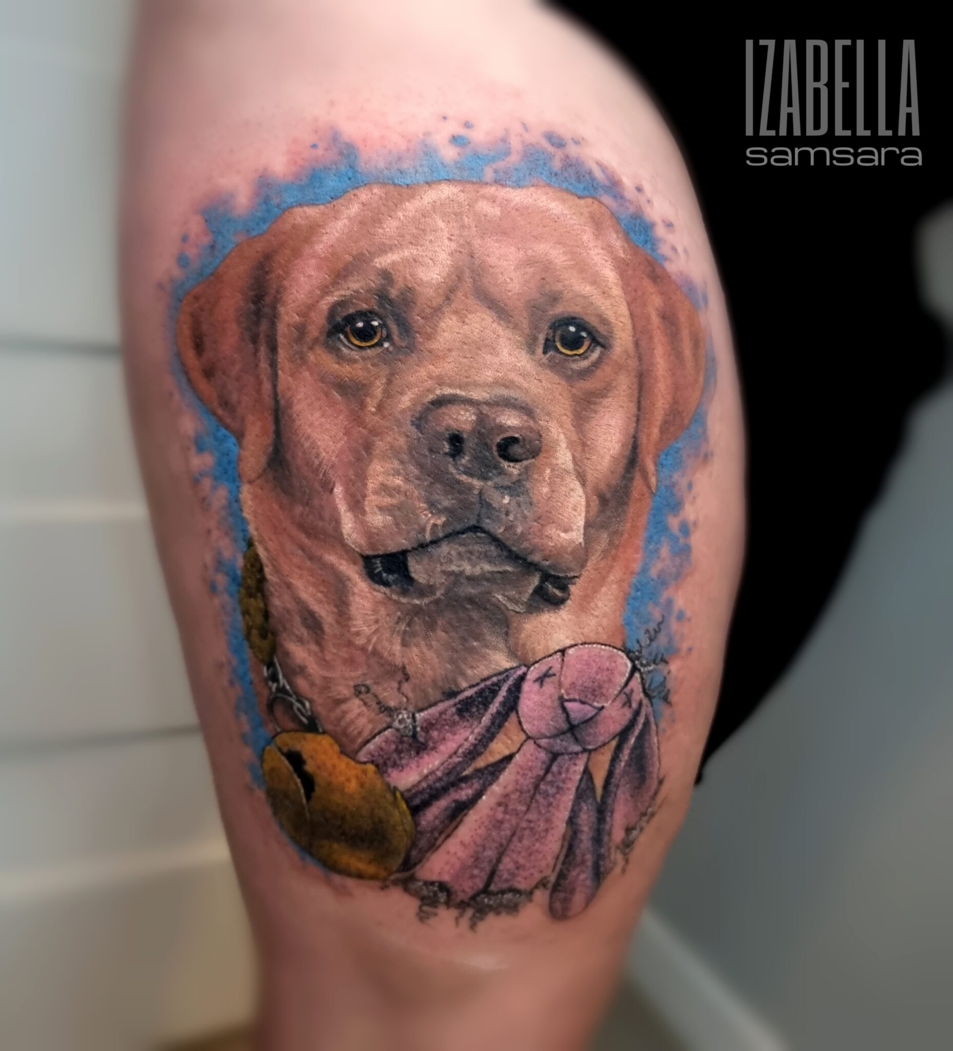A realistic tattoo of a Labrador retriever's face with a blue abstract background. The dog has a gentle expression, featuring prominent eyes, a broad muzzle, and slightly floppy ears. Additionally, there is a decorative element below the dog, resembling a pink ribbon with a bell, adding a touch of whimsy to the design.