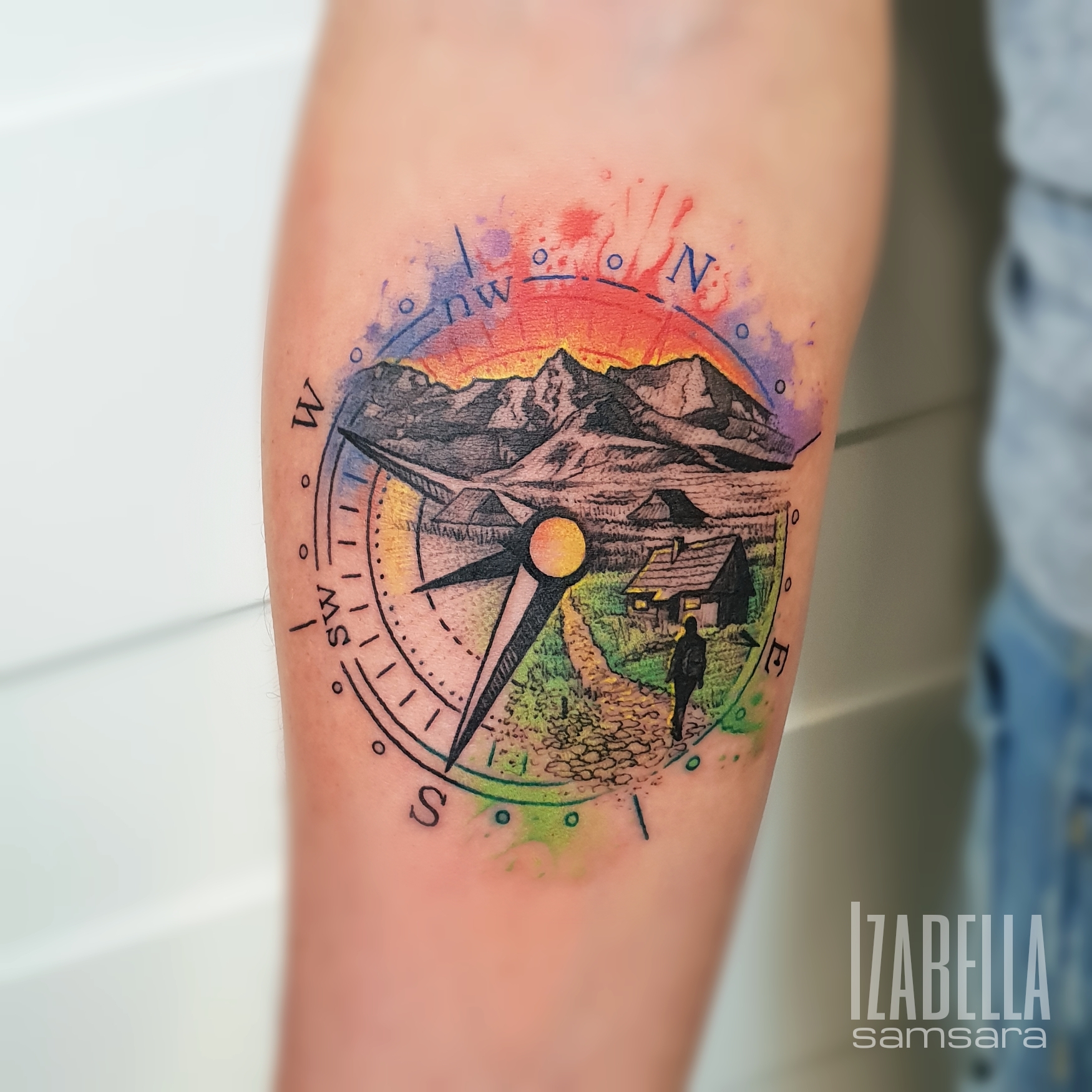 Colorful tattoo featuring a detailed compass design with a picturesque landscape. The compass, oriented with cardinal directions, is surrounded by vibrant mountains and a serene pathway, leading to a small cabin. The background showcases a gradient sunset, adding depth and warmth to the scene.