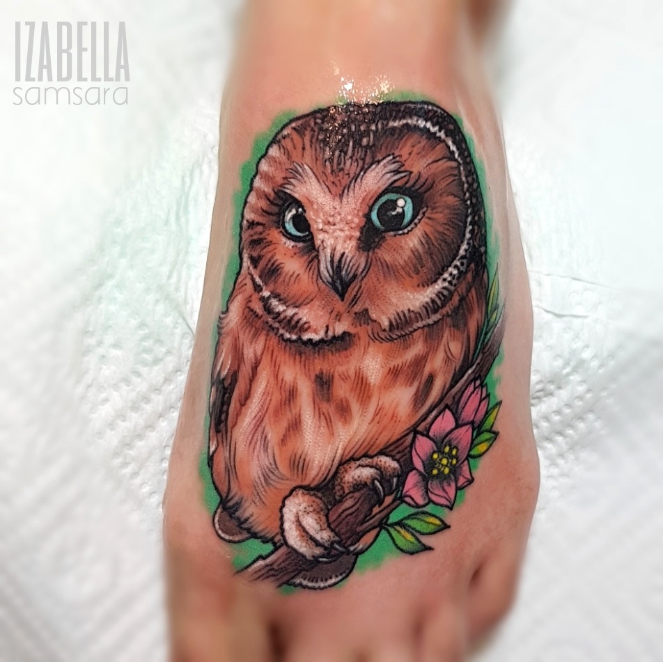 A colorful tattoo of an owl perched on a branch, featuring intricate details and vibrant colors. The owl has large, expressive blue eyes and a textured brown and white plumage. Surrounding the owl are delicate flowers in pink and green leaves, adding a natural element to the design.