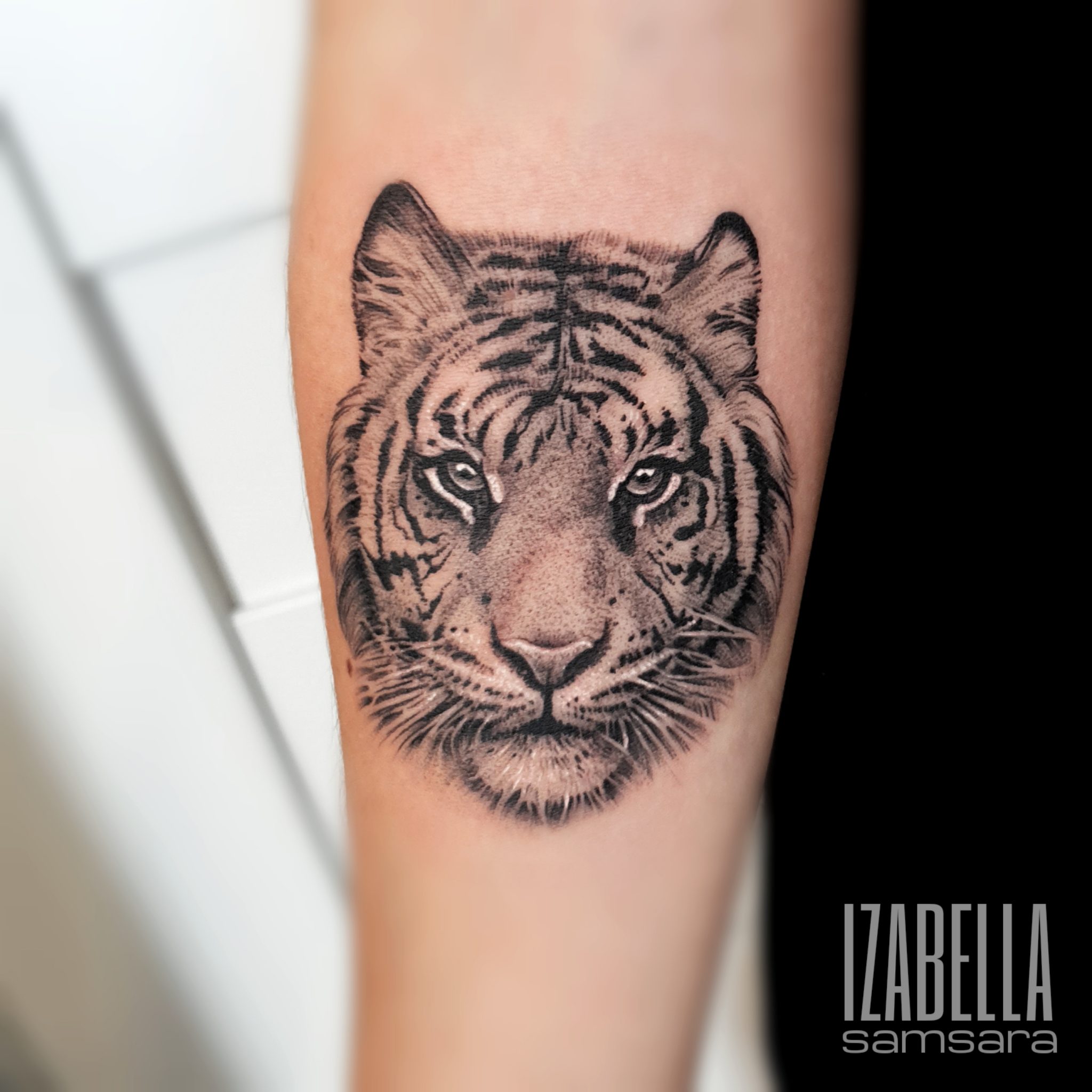 A detailed black and gray tattoo of a majestic tiger's face, showcasing intricate line work and shading. The tiger's intense eyes and pronounced stripes are vividly rendered, capturing its fierce expression. This tattoo is placed on the forearm, emphasizing the realism and artistry of the design.