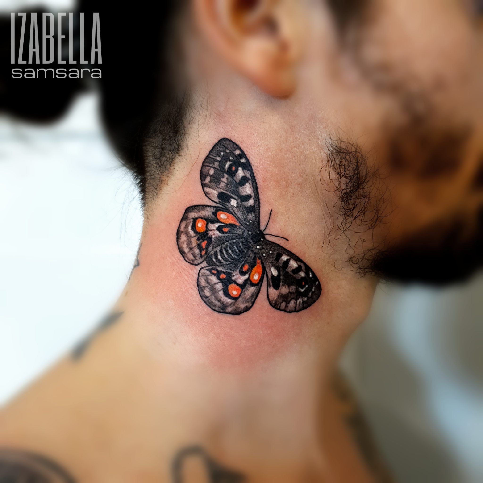 A detailed black and gray tattoo of a butterfly on the neck, featuring intricate patterns and orange accents on the wings. The butterfly is oriented with its wings spread, showcasing delicate linework and shading that adds depth and realism to the design.