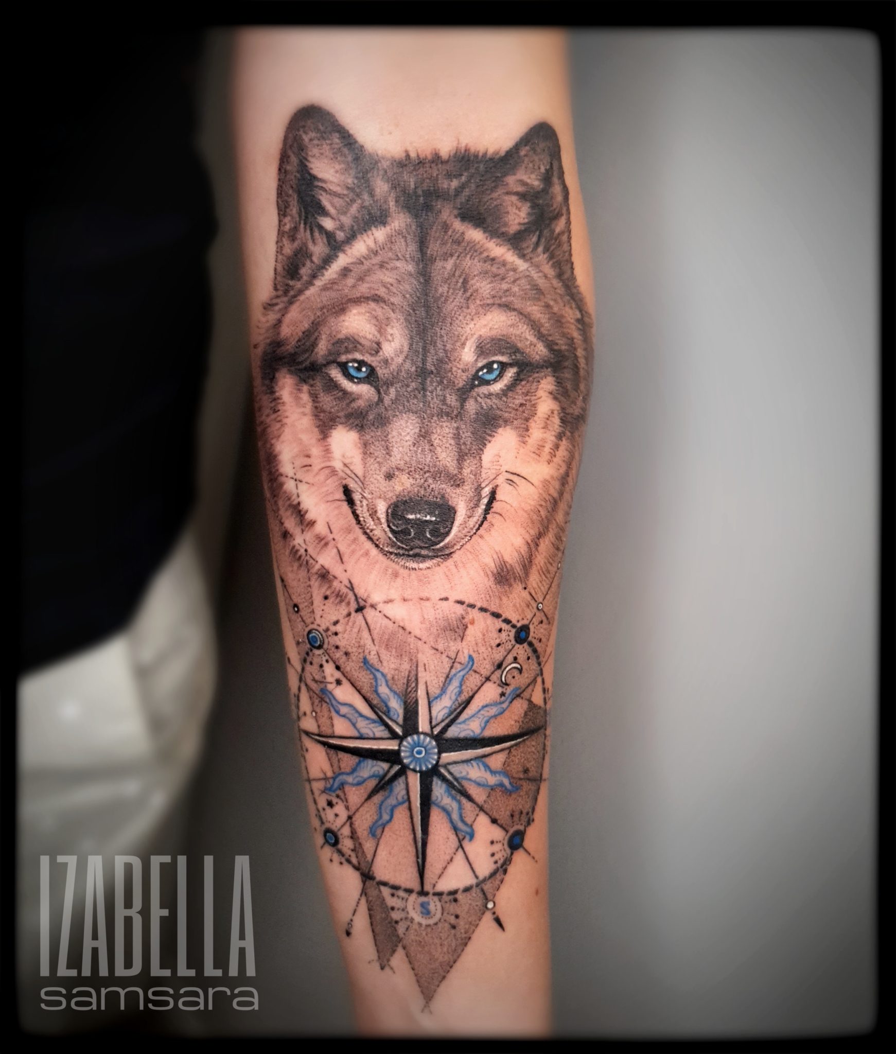 A hyper-realistic tattoo of a wolf's face with striking blue eyes, set against a geometric compass design. The wolf features detailed fur and a gentle expression, while the compass includes elegant blue and black elements, emphasizing direction and adventure. This tattoo combines natural imagery with intricate linework, making it a captivating piece.