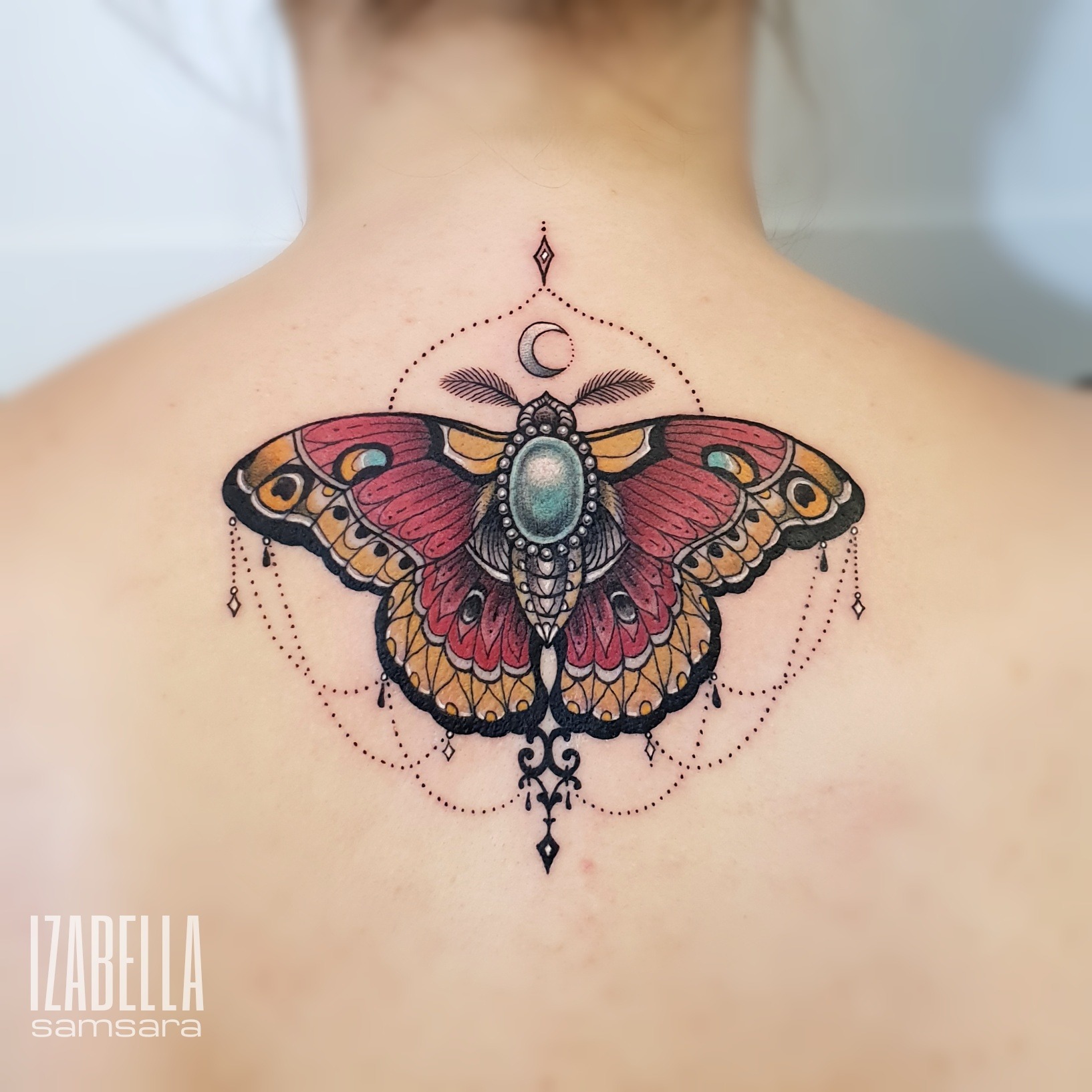 A vibrant tattoo of a butterfly with intricate details, featuring red and orange wings adorned with blue and yellow accents. The butterfly is centered on the upper back, surrounded by delicate dot work and decorative elements, including a crescent moon and dangling charms, creating a mystical and elegant appearance.