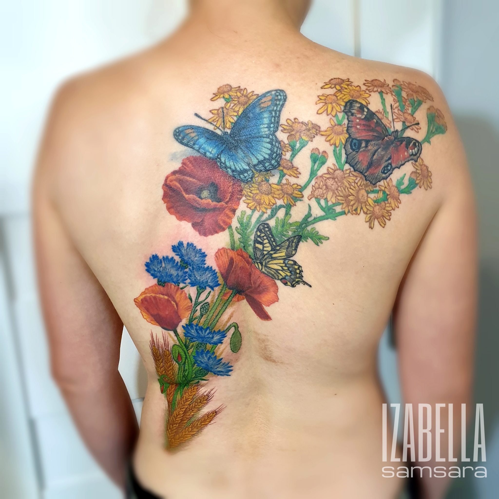 Colorful tattoo design on the back featuring a variety of floral elements including red poppies, yellow and orange flowers, and blue cornflowers. The artwork also includes multiple butterflies in vibrant colors such as blue and red, adding a lively and natural aesthetic to the tattoo.