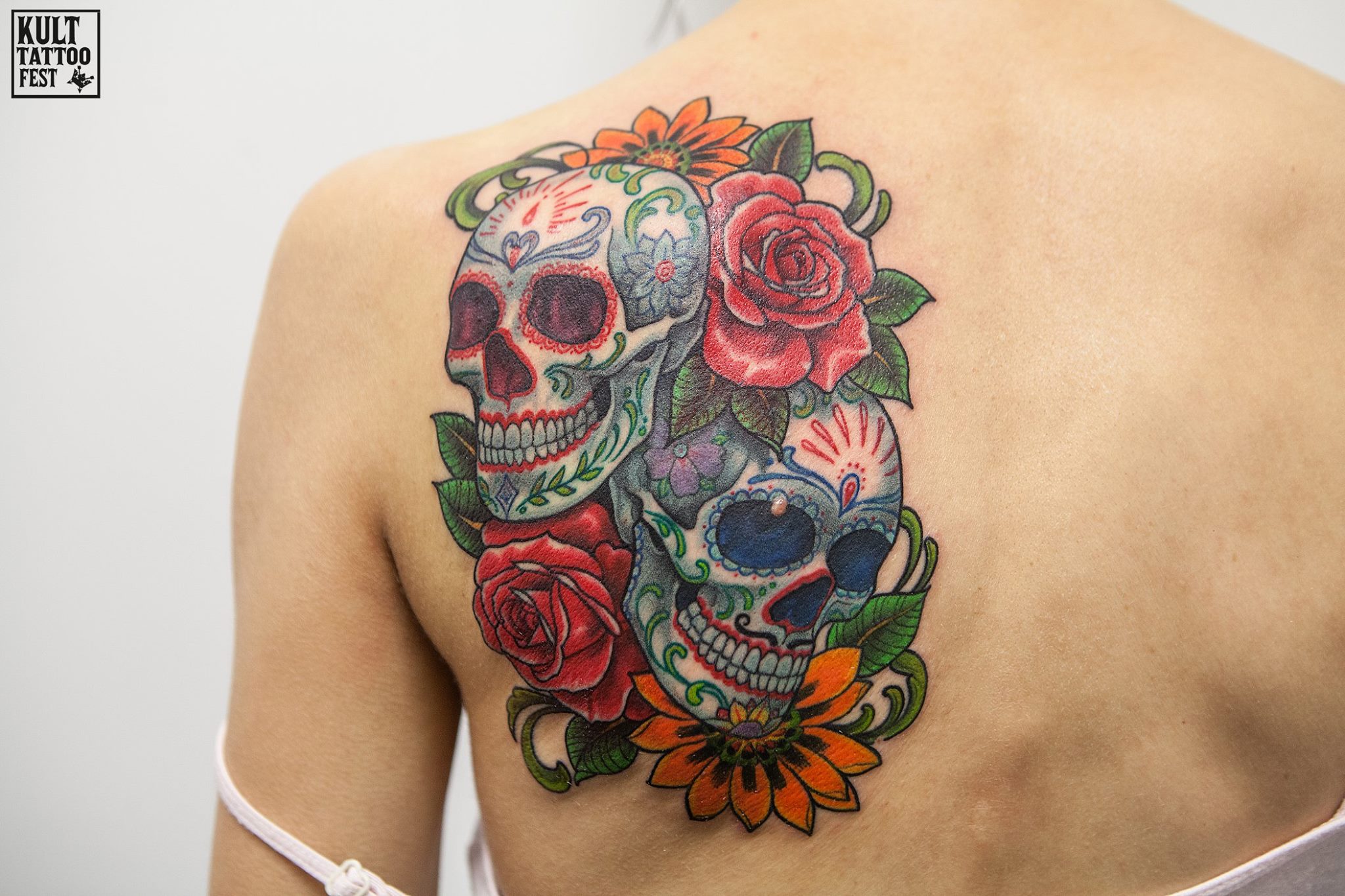 A vibrant tattoo featuring two intricately designed skulls surrounded by colorful roses, green leaves, and flowers, capturing a Day of the Dead theme with detailed patterns and vivid colors.
